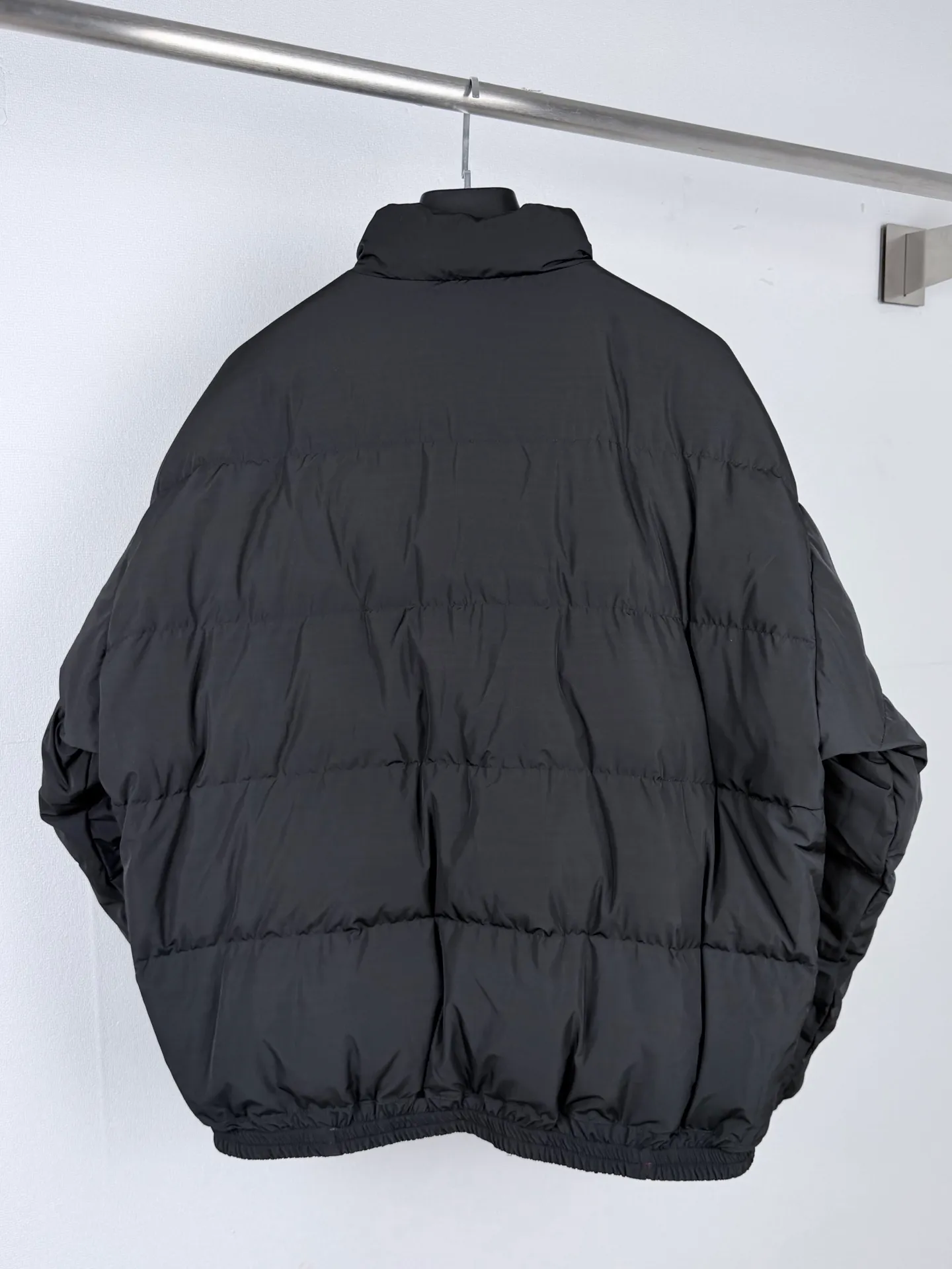 BALENCIAGA / Balenciaga 26SS black and gray reversible ring logo stand-up collar down jacket. This reversible stand-up collar down jacket combines a dual-version appearance with a 90 white duck down lining. One side presents a simple and elegant Black down bread silhouette, and the other side has gray and wine red contrasting panels for a sporty style. Both sides can be worn independently. The craftsmanship is far more complex than ordinary single-sided down garments. In the pattern making and development stage, it is necessary to complete the simultaneous calibration of the two plates. The shoulder slope, armhole arc, zipper placket, and hem elastic point of the front and back sides must be perfectly matched. Once there is a slight deviation in the positioning of the cutting pieces, it will lead to misalignment of the pattern and unbalanced length of the garment when worn on both sides. The down side adopts a horizontal segmented compartment design and is filled with high-quality 90 white duck down. The amount of down filling in each down compartment is accurately weighed and distributed to ensure that the down is evenly distributed and avoid local accumulation or gaps of down material. The fabric is made of high-density anti-drilling down base to minimize the risk of down escaping. The brand logo on the left chest adopts fine thermal transfer printing technology, and the positioning and color number have been reviewed and debugged many times. The body is equipped with a special two-way zipper on both sides, which can be opened and closed smoothly on both sides. The placket adopts a sandwich hidden hemming process, and all sewing lines are stored on the inside of the sandwich. There are no unnecessary exposed stitches on both sides, ensuring a clean and neat appearance. The welt pockets on both sides have an interconnected double-sided structure. The three layers of fabric are aligned simultaneously during sewing. The pocket opening cannot leave stitches on either side that would damage the look and feel. The cuffs and hem adopt hidden elastic hemming technology, and the elastic bands are completely wrapped in the interlayer. There are no traces of elastic bands on the front and back, taking into account windproofness, temperature locking and beautiful lines. The sewing production process is extremely difficult. The sewing operation requires penetrating three layers of outer fabric, 90 fleece liner, and reverse woven fabric at one time. Turners have extremely high requirements for controlling the tightness of stitches. If the stitching is too heavy, it will compress the down bin, damage the down structure, and reduce the warmth retention performance; if the stitching is too loose, it is easy to cause jumpers and down drilling problems. Multiple special-shaped pieces in the contrasting area on the back are spliced ​​with White welts, and the dividing seams must be accurately aligned with the Black side seams. After the garment is sewn, the two facades need to be shaped and ironed separately. One side is baked to give the fluffy and full texture of the down, and the other side is calibrated to make the contrasting color dividing lines straight. The entire production process implements double quality inspection standards, which takes longer, and ultimately allows one piece of clothing to unlock two styles. Size:XS S M L